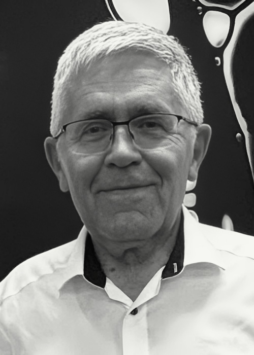 Older man with gray hair and glasses, wearing a white shirt, smiling at the camera in a black-and-white portrait.