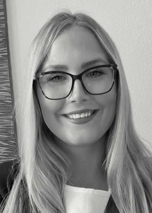 Black-and-white portrait of a smiling woman with long blond hair, wearing dark-framed glasses and a blazer indoors.